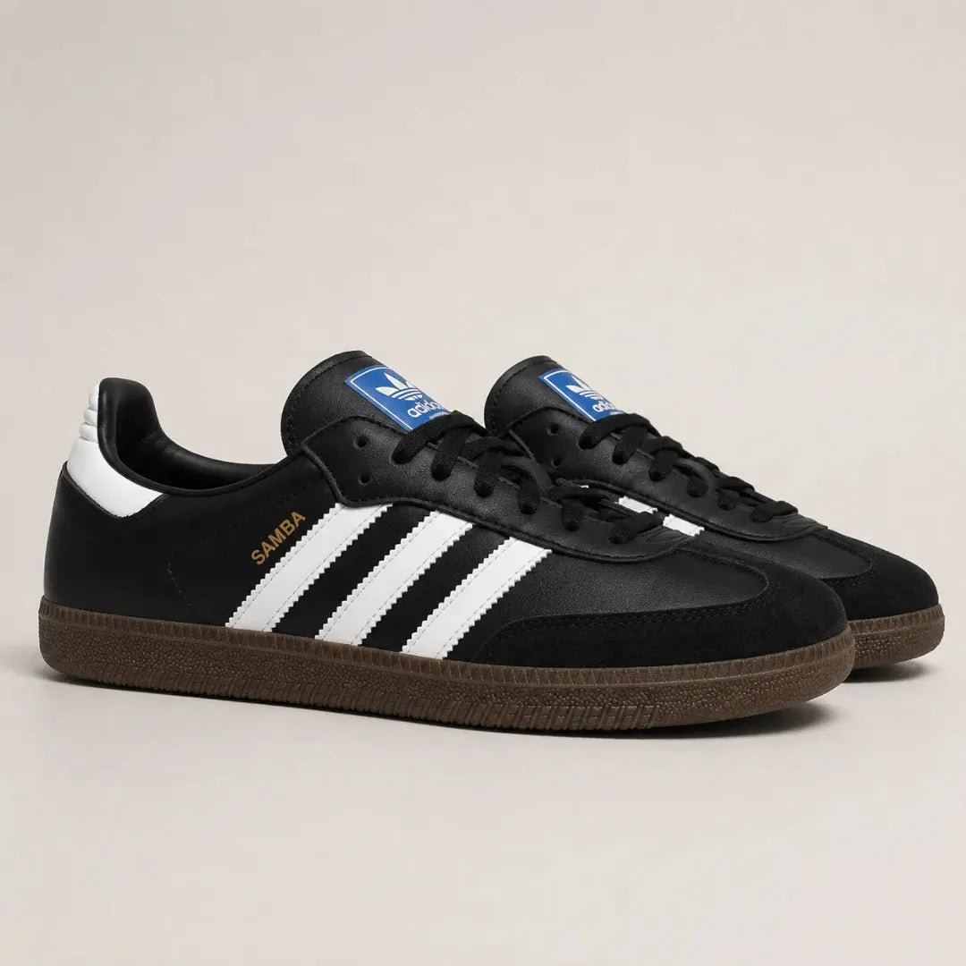 A pair of Adidas Samba sneakers in black, featuring three white stripes, a blue tongue label, gold 'SAMBA' text, and a brown gum sole, set against a plain light beige background.