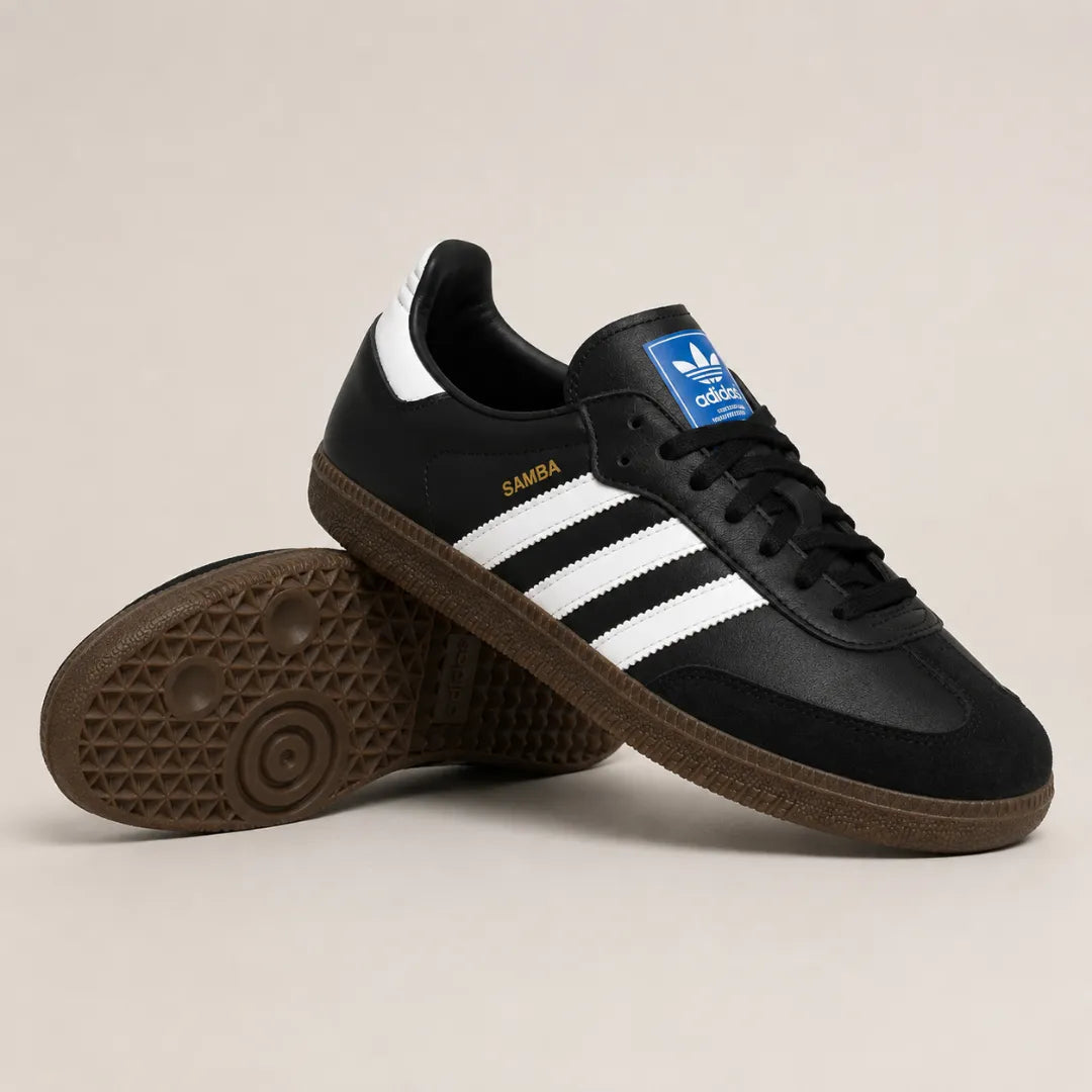 A pair of Adidas Samba sneakers in black, featuring white three-stripes on the side, a blue Adidas logo patch on the tongue, and a brown gum sole, displayed on a light beige background.