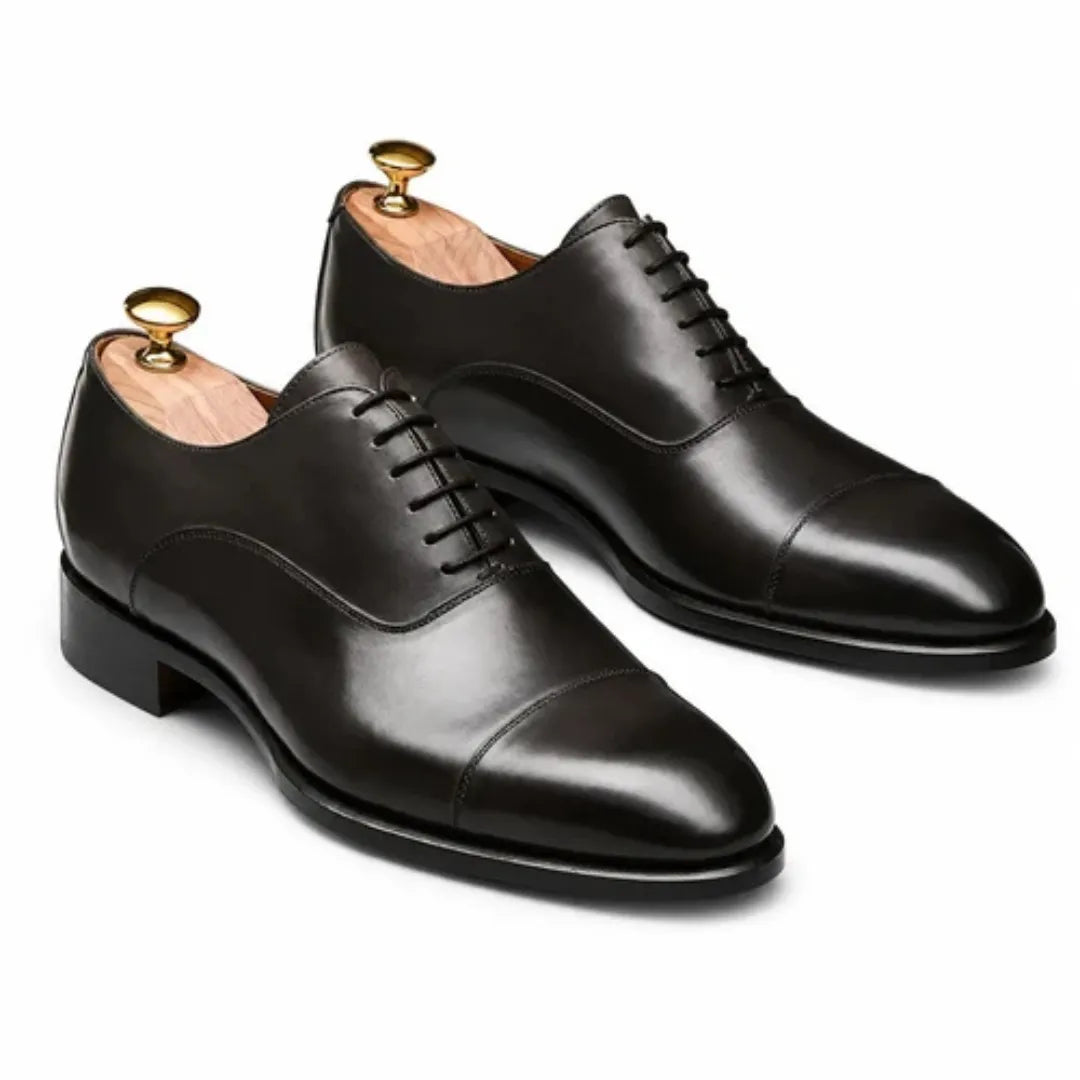 A pair of black leather cap-toe oxford dress shoes with black laces, displayed on wooden shoe trees with gold tips against a clean white background.